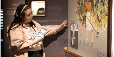 Mujer usando audifonos y con un celular en su mano señalando un cuadro en la pared.