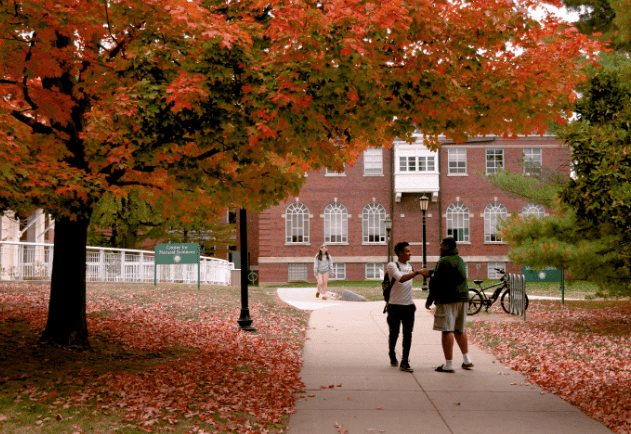 Paisaje de otoño, dos personas caminando, arboles con hojas naranja y uno de los edificios de IWU en el fondo.