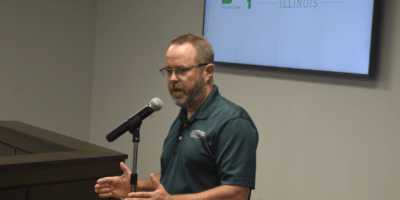 Matt Burgess, director ejecutivo de Home Sweet Home Ministries, de pie frente a un podio durante una reunión del Consejo del Condado de McLean, gesticulando mientras responde preguntas sobre el proyecto del refugio The Bridge.