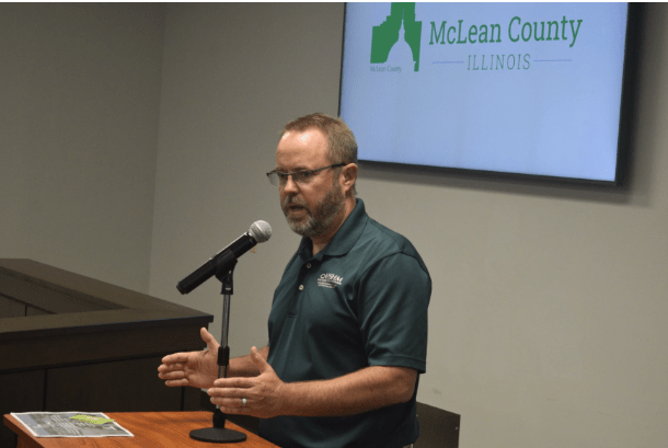 Matt Burgess, director ejecutivo de Home Sweet Home Ministries, de pie frente a un podio durante una reunión del Consejo del Condado de McLean, gesticulando mientras responde preguntas sobre el proyecto del refugio The Bridge.
