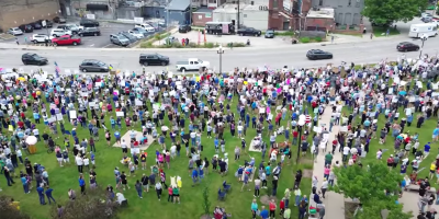 Carros circulando por una avenida y mucha gente observandolos y gritando consignas.