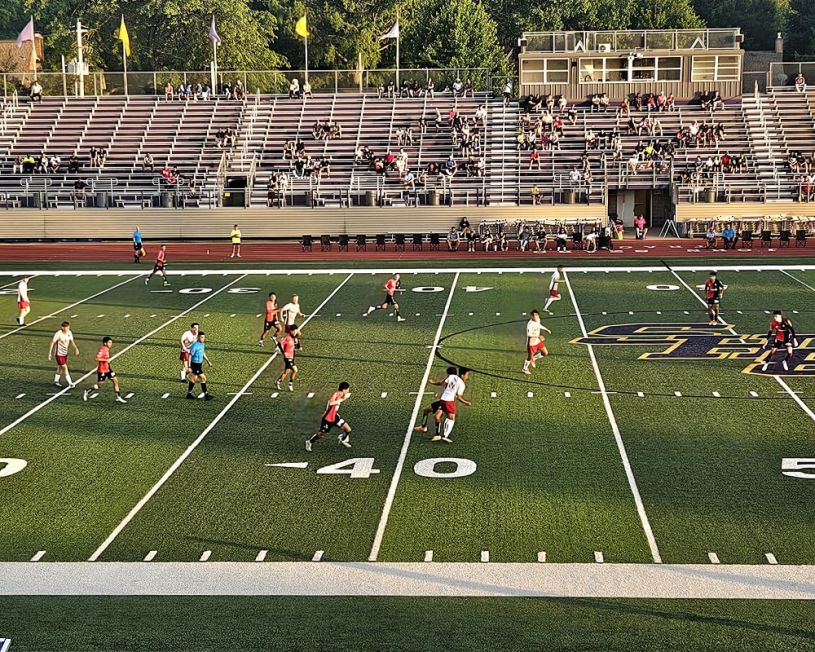 Campo de futbol con algunos jugadores corriendo y el publico sentado en las bancas de fondo