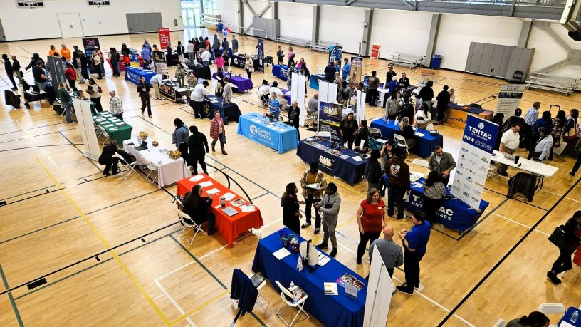 Vista aérea del gimnasio del YMCA con mesas de empresas y personas interactuando en la feria de empleo.