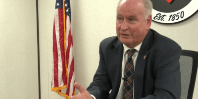 Alcalde Dan Brady sentado en su escritorio con el sello de la ciudad detras y la bandera de USA a un lado.