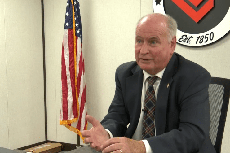 Alcalde Dan Brady sentado en su escritorio con el sello de la ciudad detras y la bandera de USA a un lado.