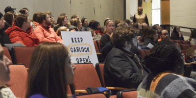 Miembros de la Junta Escolar de Unit 5 sentados durante una reunión pública en Bloomington mientras residentes observan y participan en comentarios comunitarios.