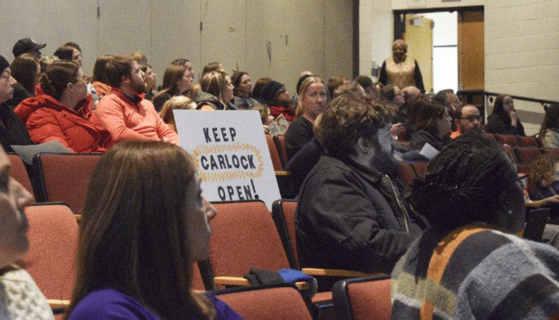 Miembros de la Junta Escolar de Unit 5 sentados durante una reunión pública en Bloomington mientras residentes observan y participan en comentarios comunitarios.