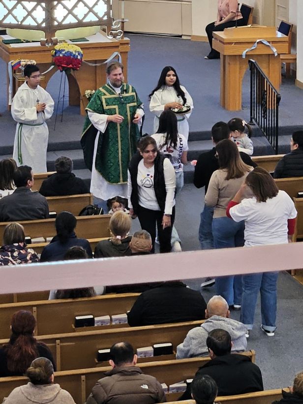 una fila de 5 personas en el centro de la iglesia, la primera entrega una ofrenda y el padre la recibe.